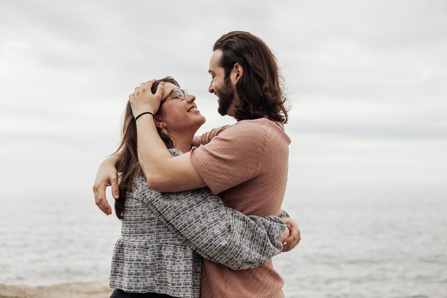 Romantic couple embracing and smiling at each other by the ocean on a cloudy day.