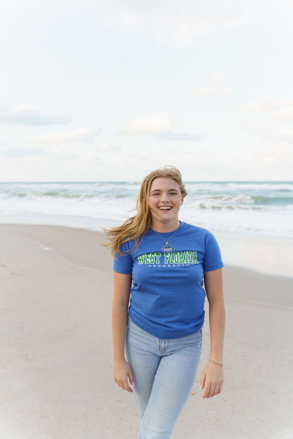 Smiling young woman in blue West Florida t-shirt and jeans standing on a sandy beach with ocean waves behind her.