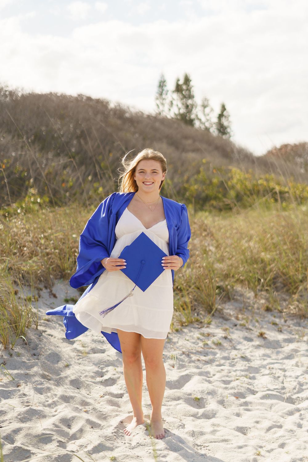 Smiling graduate in blue cap and gown on sandy beach dunes, holding mortarboard, wearing white dress.