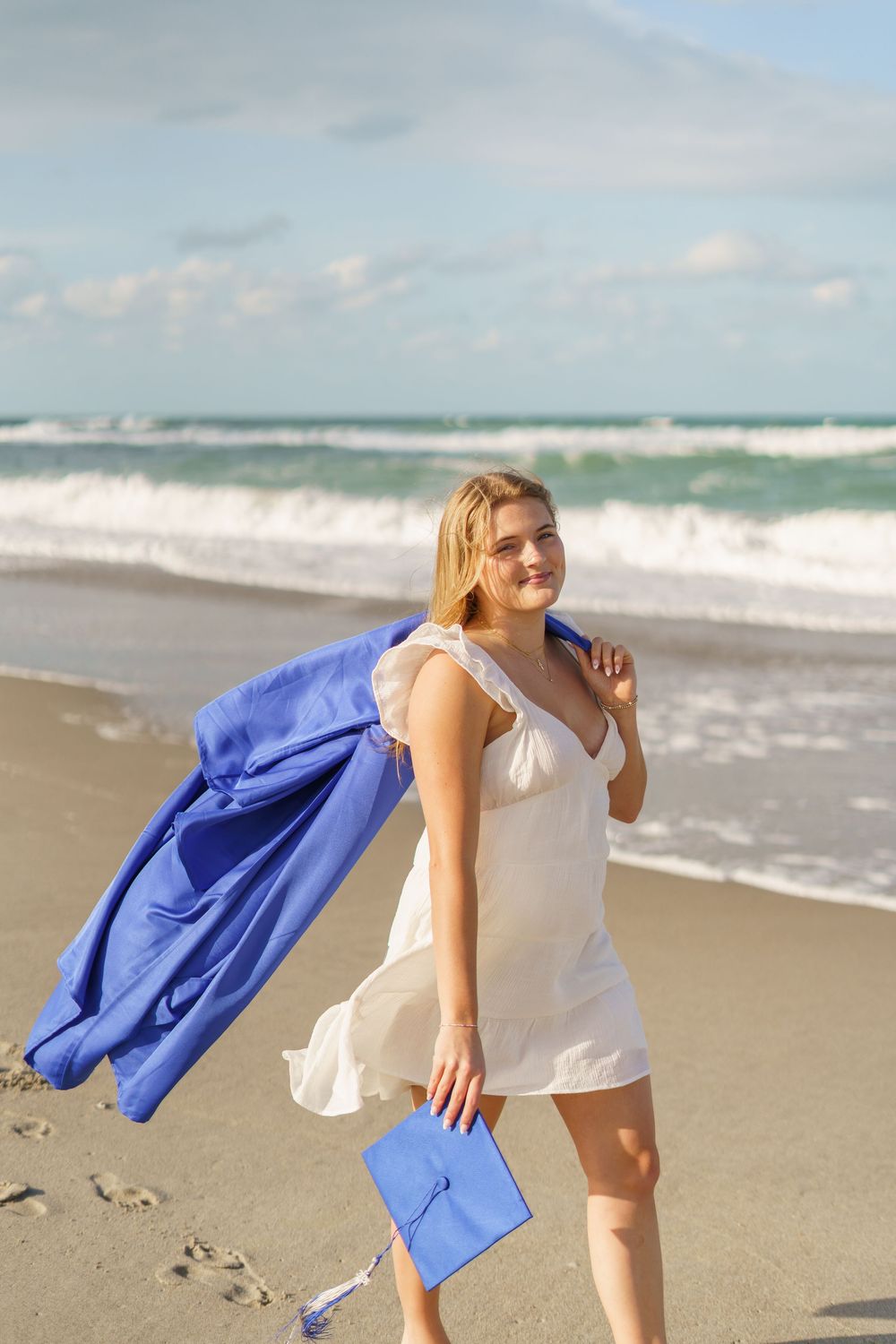 Smiling blonde graduate in white dress holding blue cap and gown on a sunny beach.
