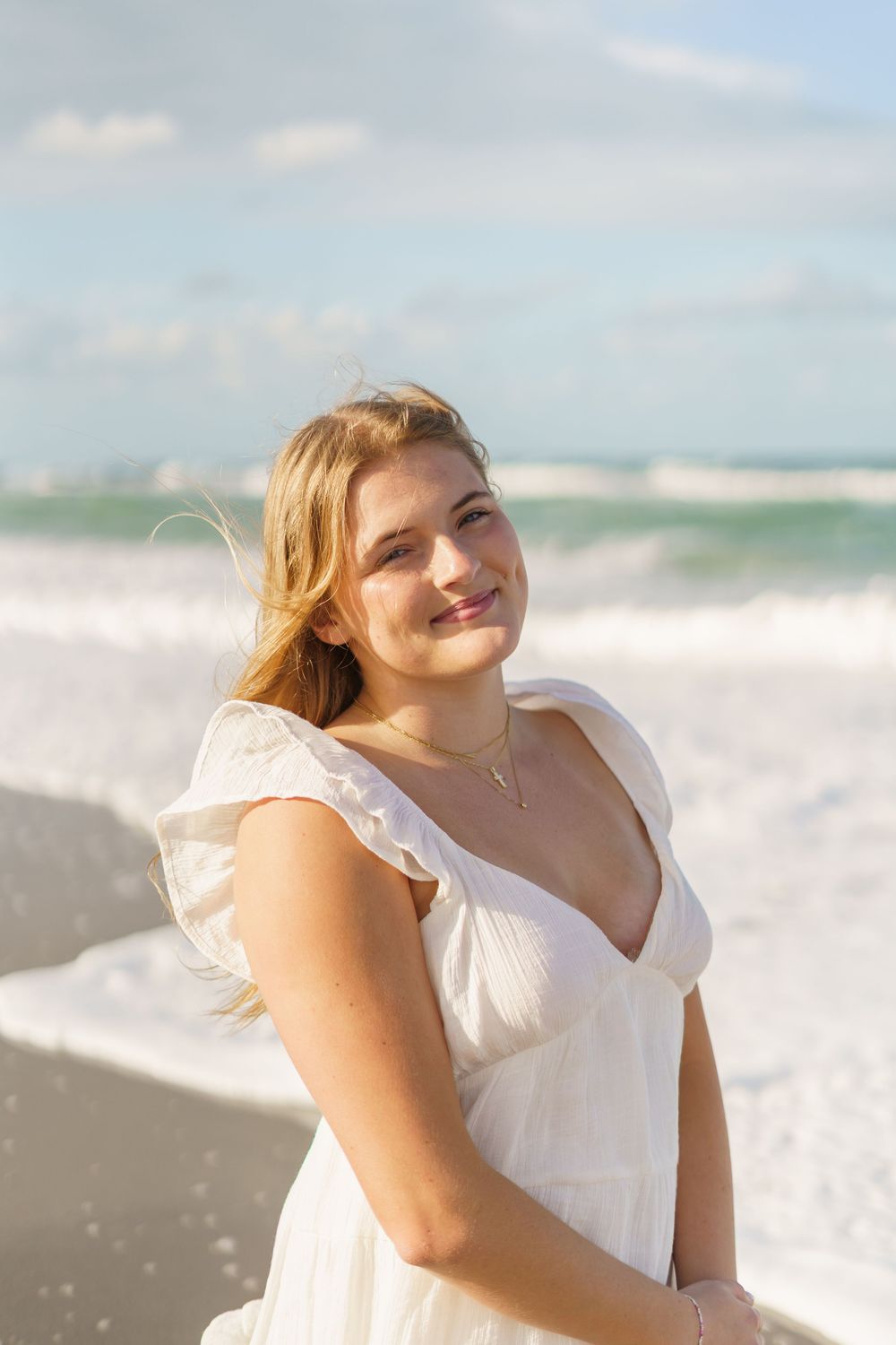 Young blonde woman in white ruffle dress smiling at beach with ocean waves behind her.