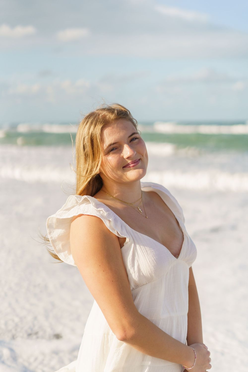 Blonde woman in white sundress smiling on a bright sandy beach with ocean in background.