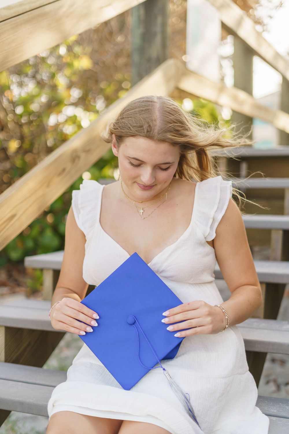 Young woman in white dress holding blue graduation cap while sitting on wooden steps outdoors.
