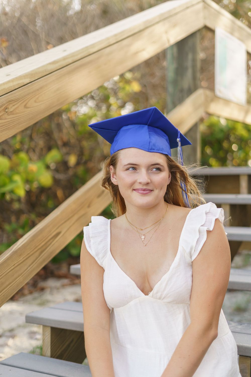Young woman in white dress wearing blue graduation cap seated on wooden steps outdoors.
