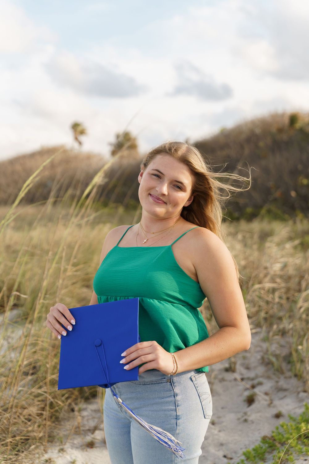 Smiling young woman in green top holding blue graduation cap on a beach dune path.
