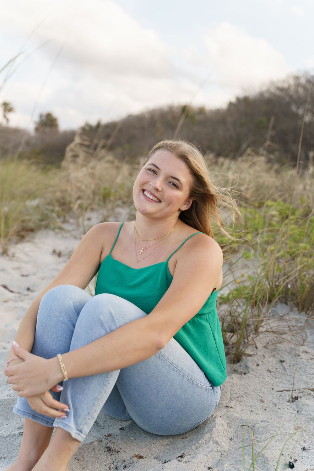Smiling young woman in green tank top and jeans sitting on sandy beach dunes with grass and cloudy sky.