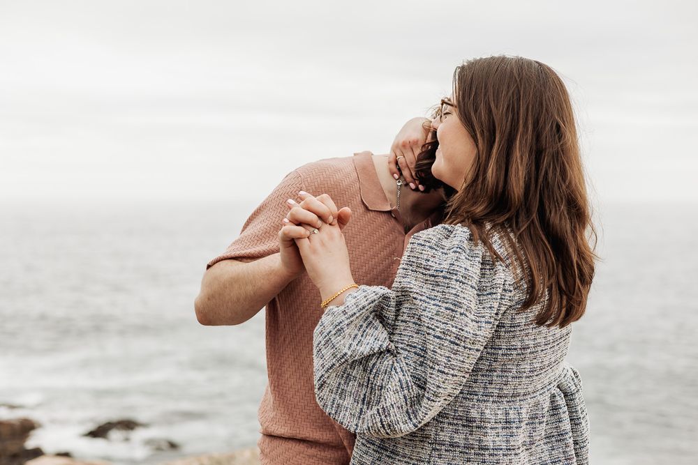 Romantic couple holding hands and embracing near the ocean on a cloudy day, sharing a tender moment.