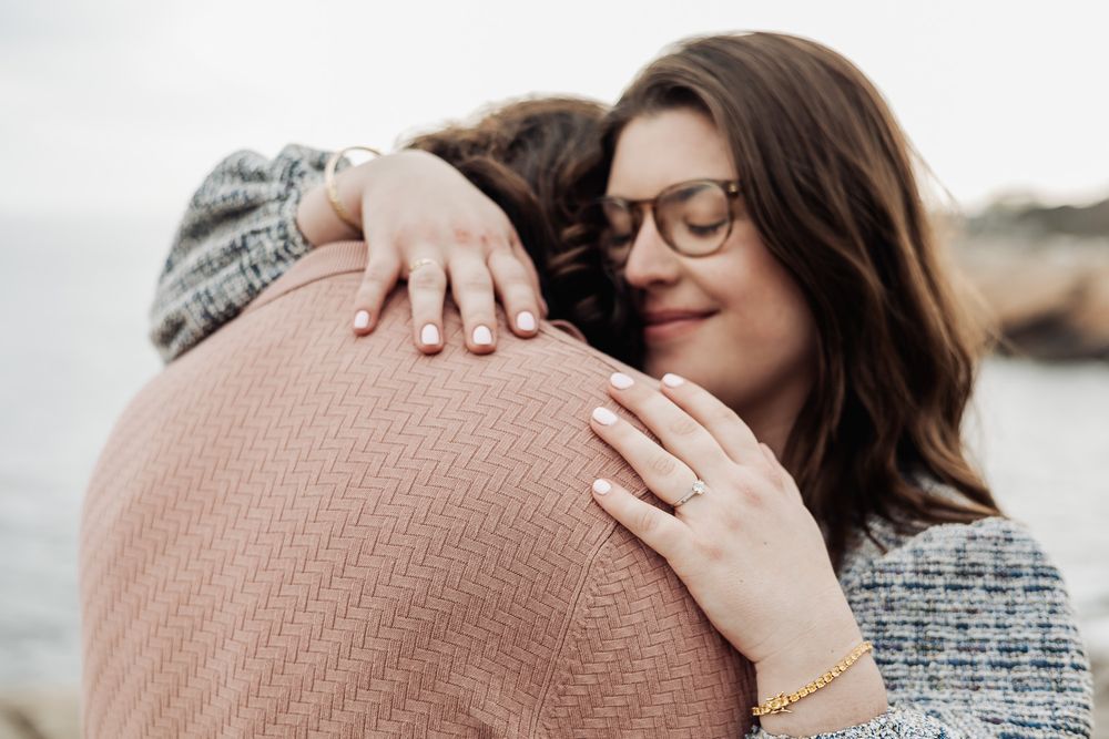 Woman with glasses smiling while hugging partner, showing engagement ring on her hand.