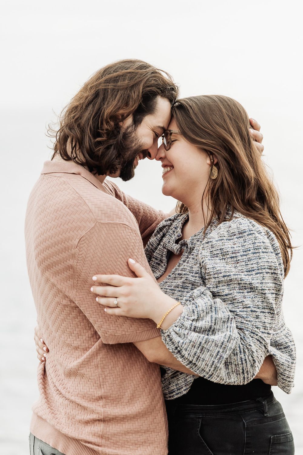 A couple smiling and embracing forehead-to-forehead near the ocean.