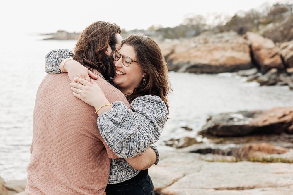 A smiling woman hugging a man near rocky coastal shoreline on a cloudy day.