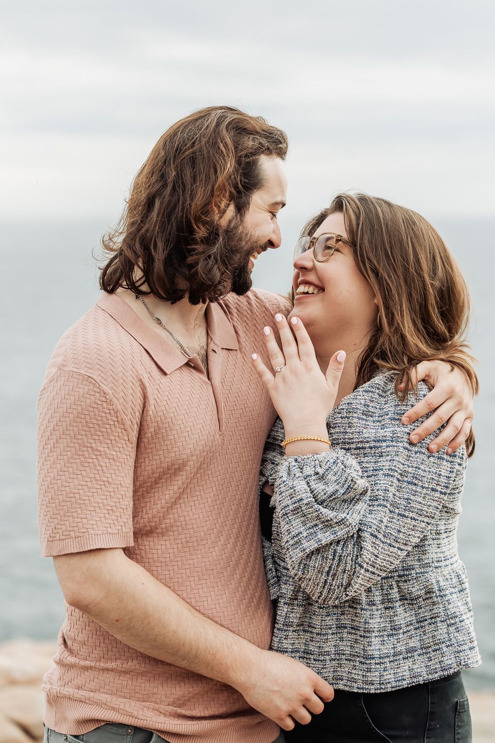 Joyful engaged couple embracing and laughing by the ocean, woman displaying ring.