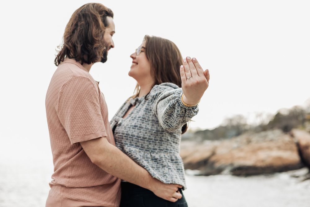 Happy couple laughing together outdoors near rocky shore, woman showing engagement ring.