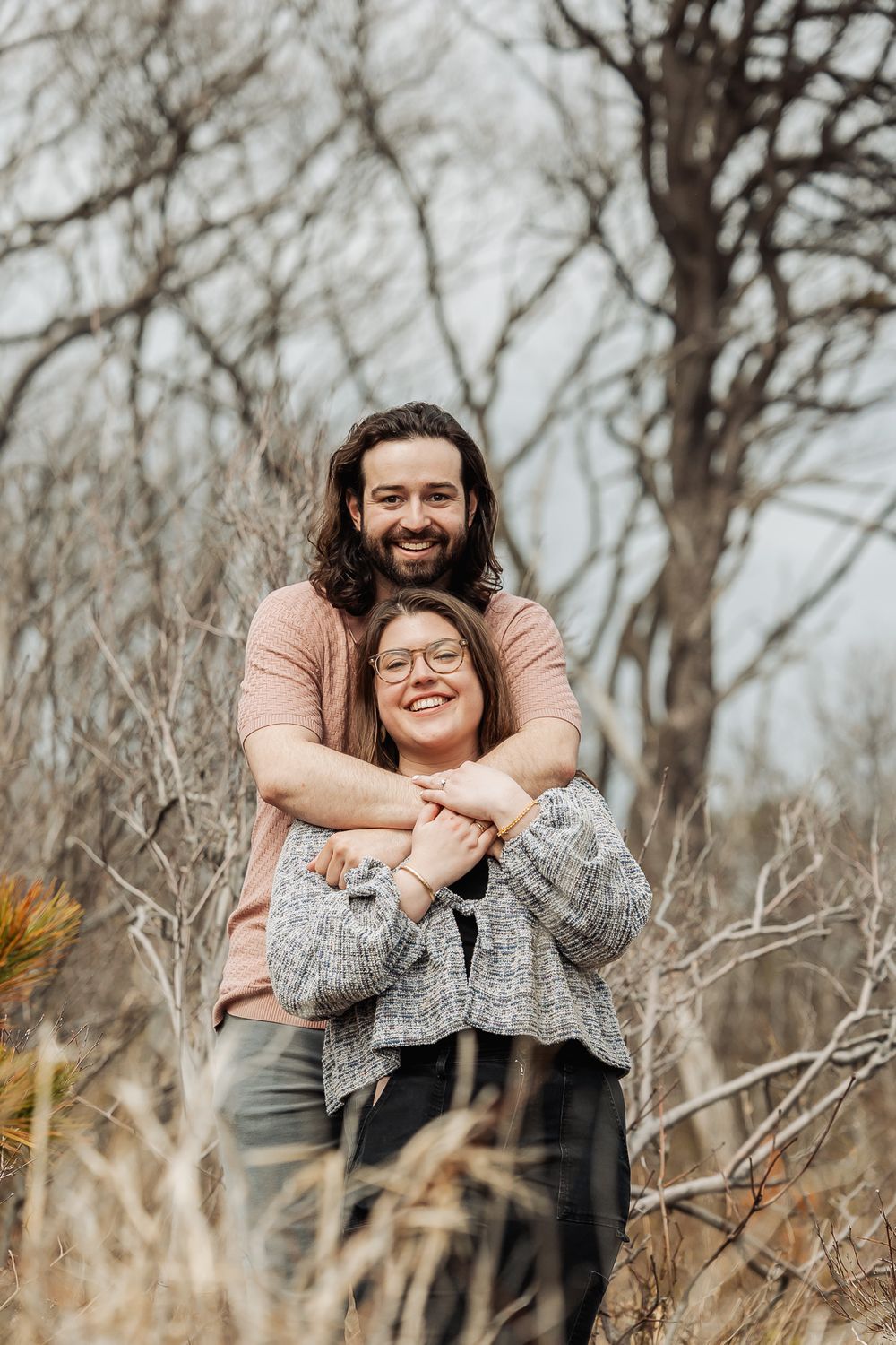 Happy couple embracing in a winter forest, man hugging woman from behind as she smiles wearing glasses and gray jacket.