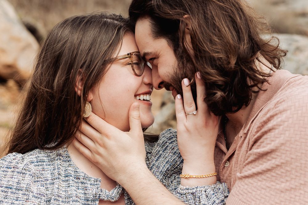 Engaged couple touching foreheads and smiling, woman showing diamond ring while man gently holds her face outdoors.