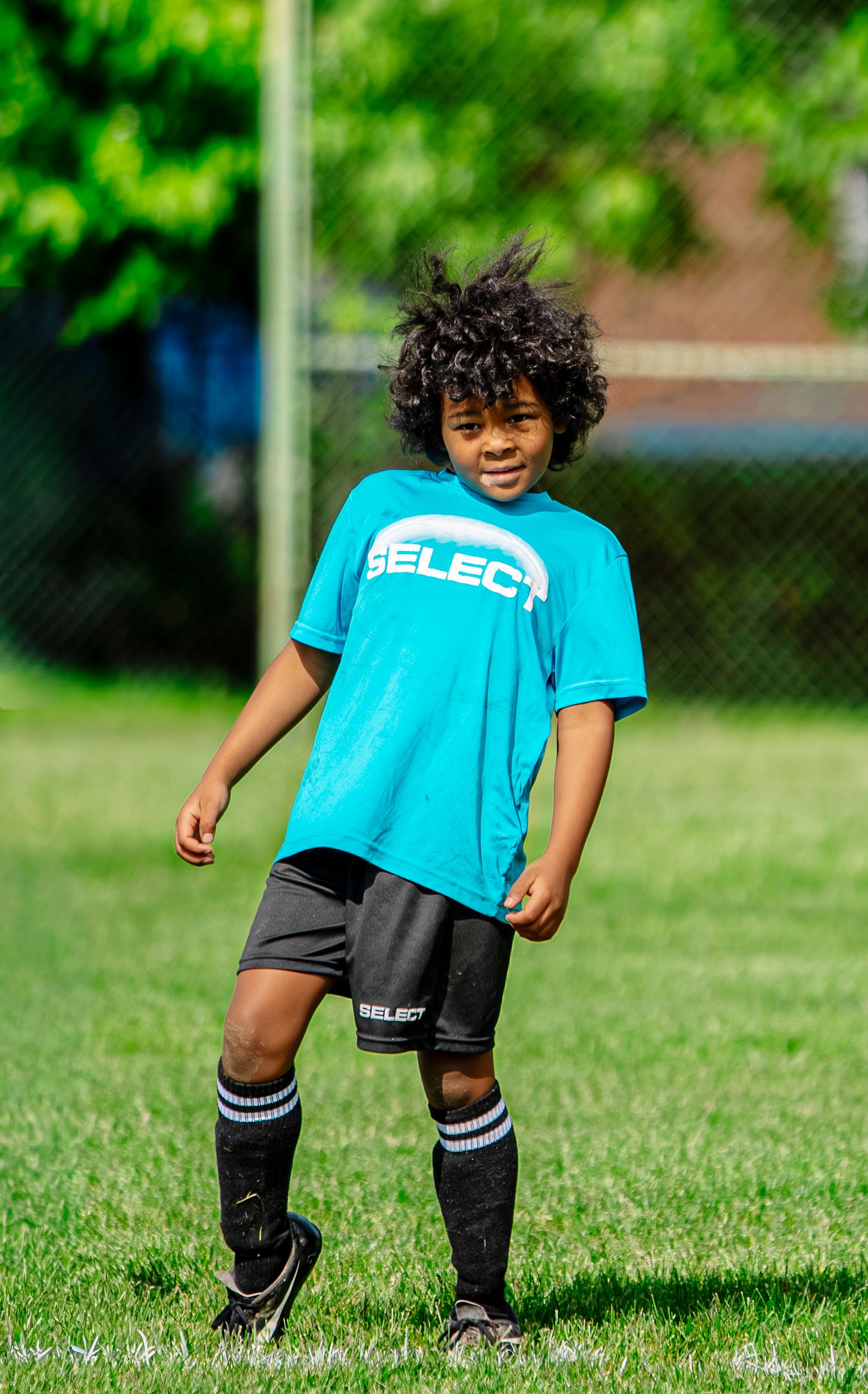 Young boy with curly hair wearing a blue SELECT t-shirt standing outdoors in a park.