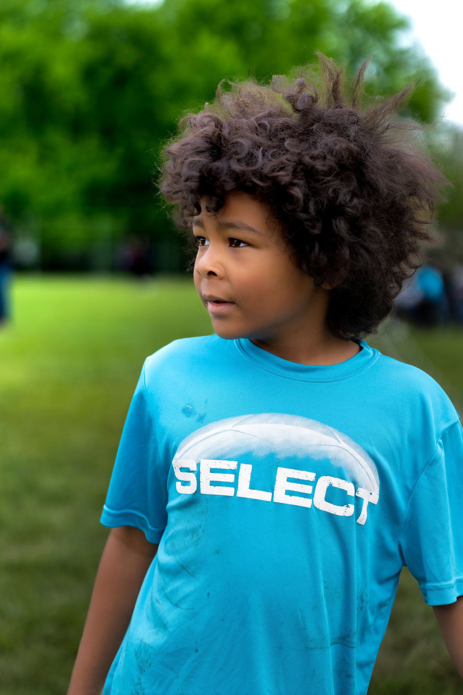 Young boy with curly hair wearing a blue SELECT t-shirt standing outdoors in a park.