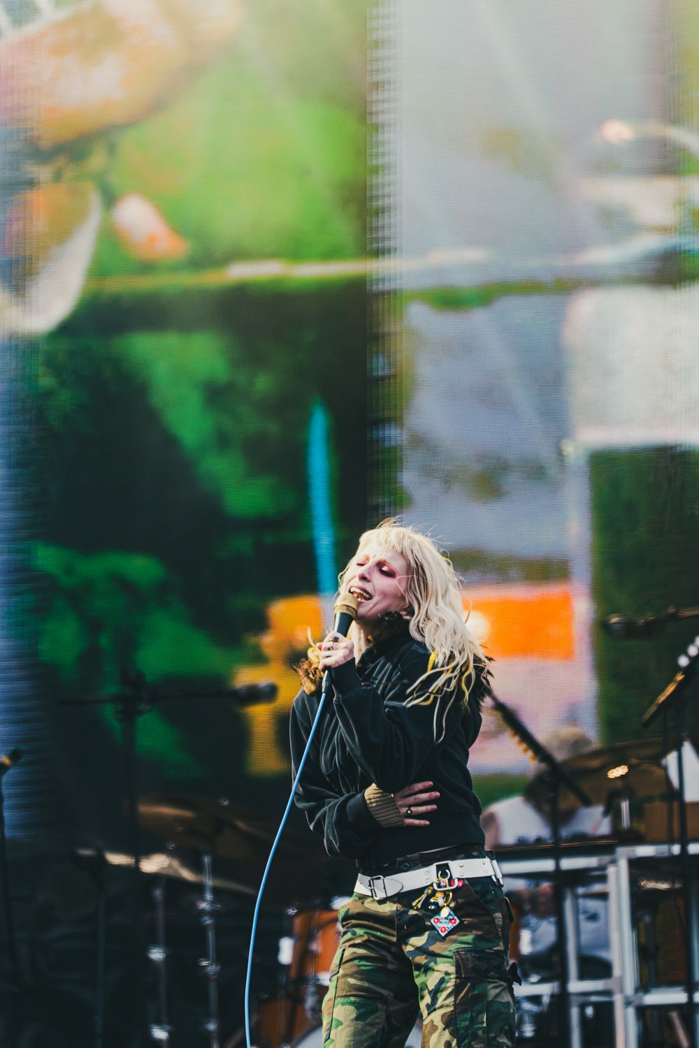 Blonde female singer performing on stage wearing camo pants and black jacket while holding microphone at outdoor concert.