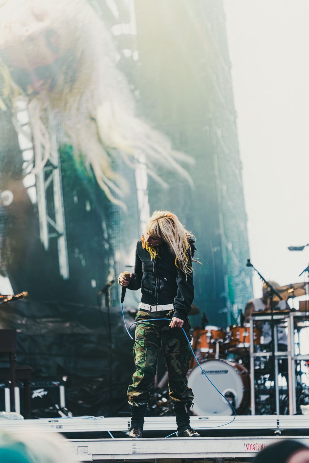 Rock performer with long blonde hair in camo pants and black jacket singing on stage at an outdoor music festival.