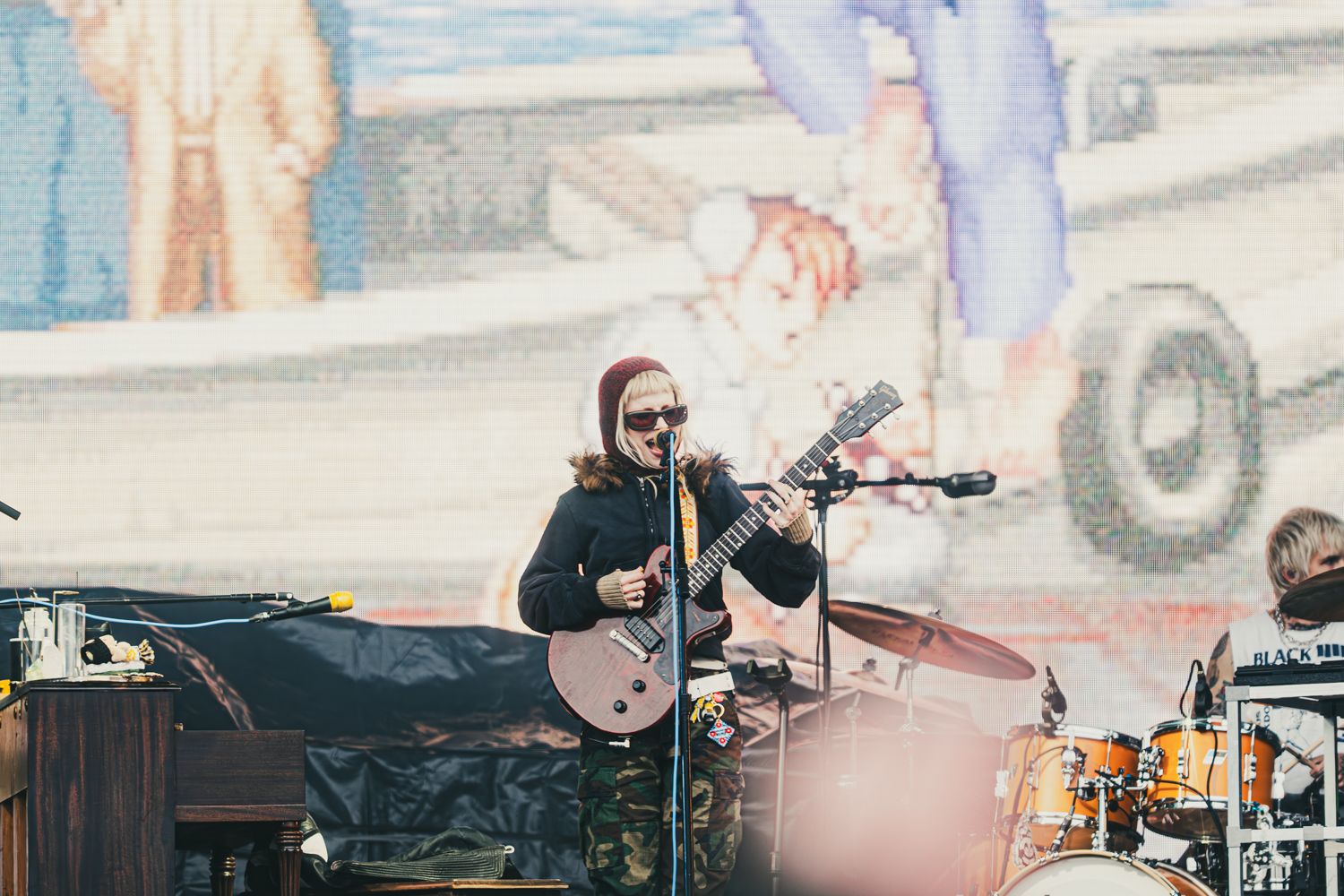 Guitarist performing on outdoor stage with colorful mural backdrop at a live music concert.