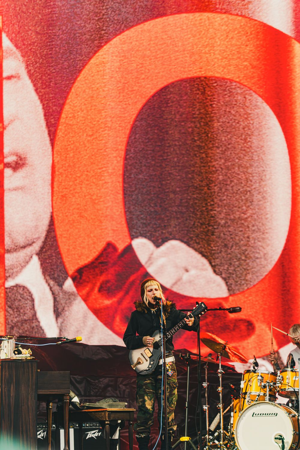 Rock musician playing guitar on stage with dramatic red backdrop featuring large circular design at outdoor festival.