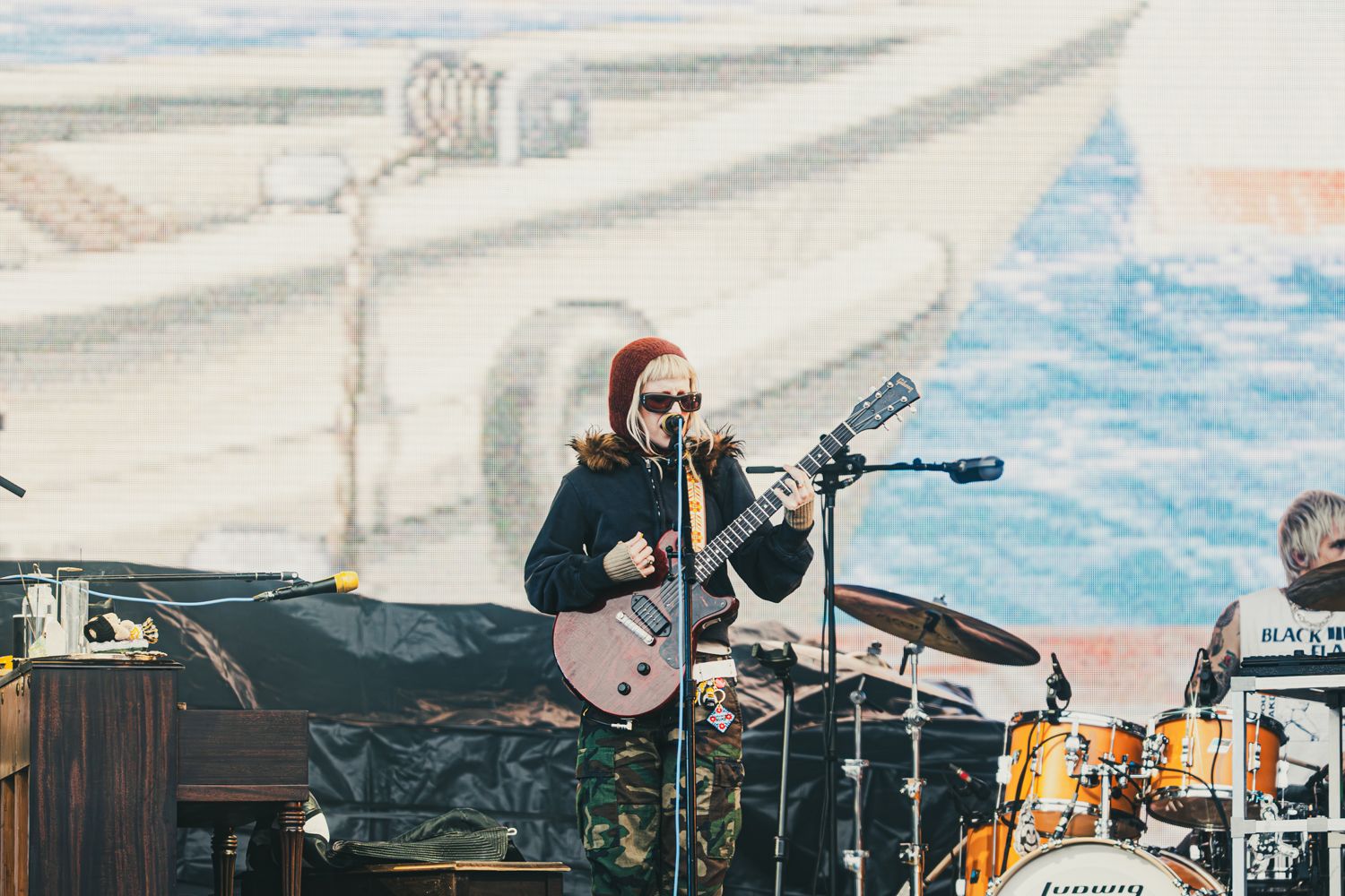 Musician playing electric guitar on outdoor stage with colorful mural backdrop at live concert.