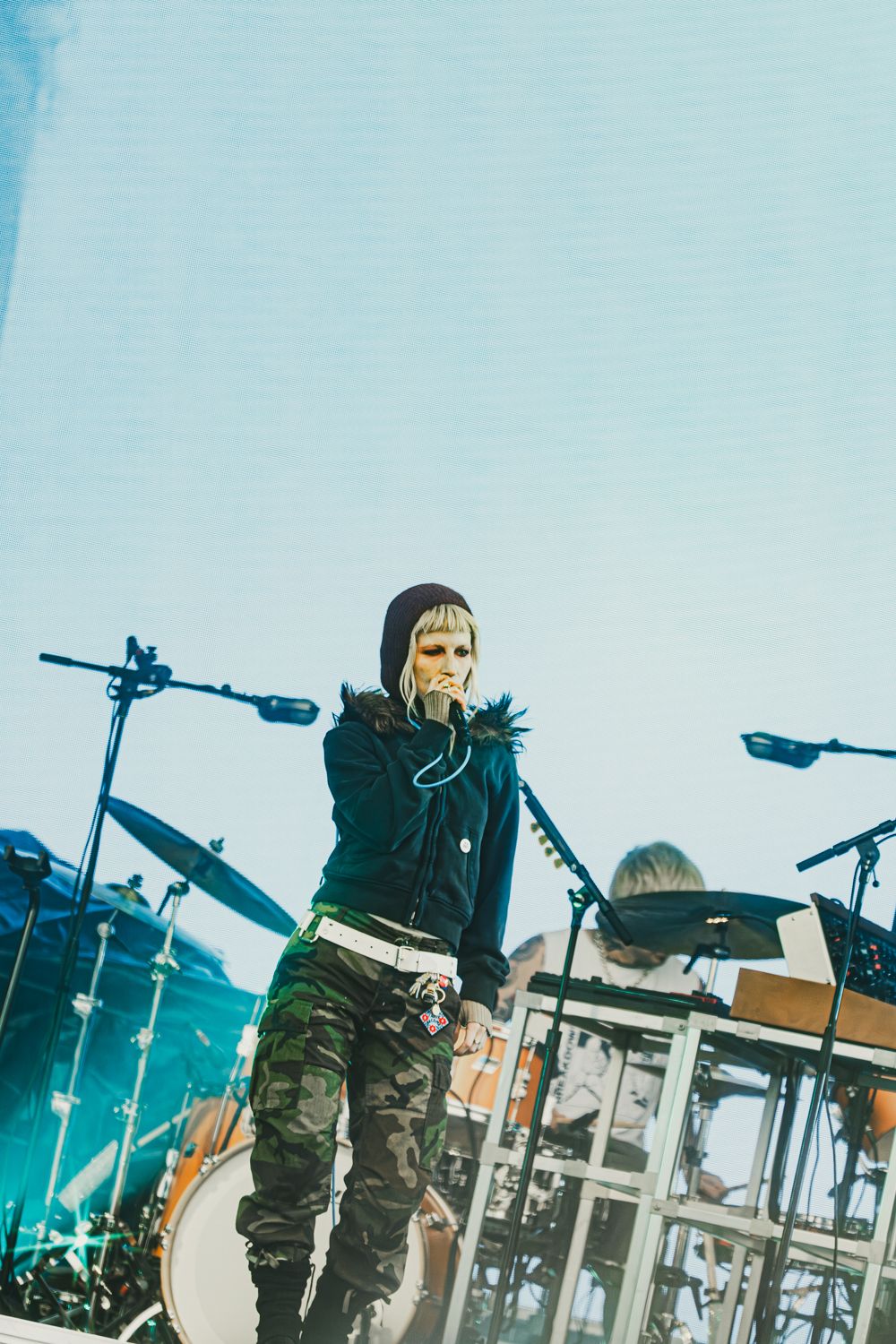 Singer in camo pants and black hoodie performs on outdoor stage with drums in background.