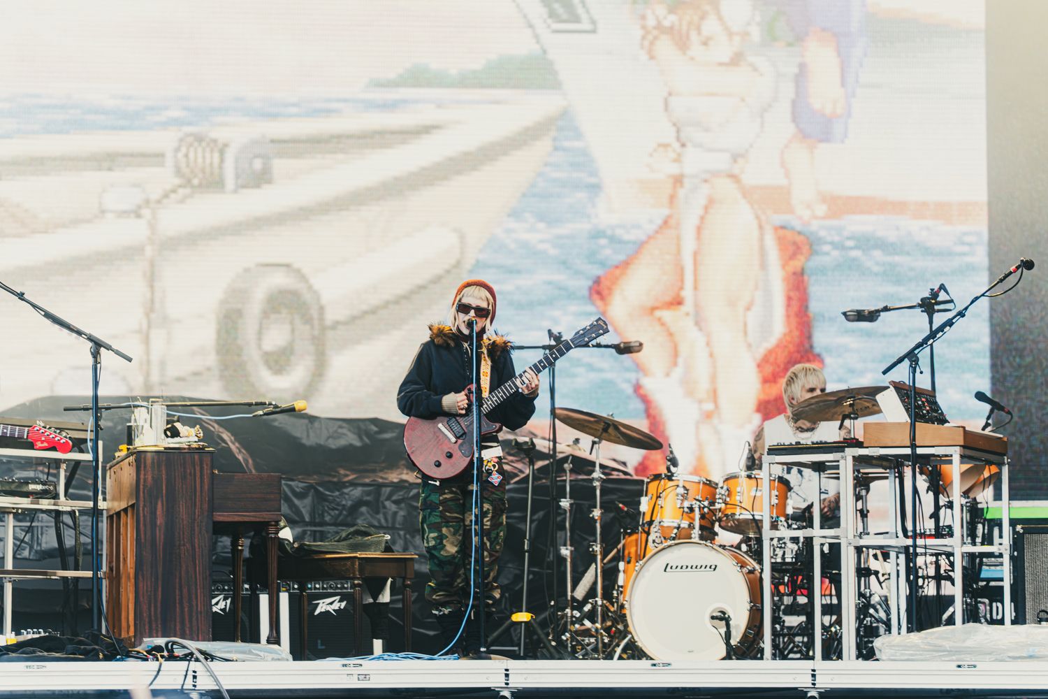 Musician playing guitar on outdoor stage with vintage beach mural backdrop and drum kit during live concert.