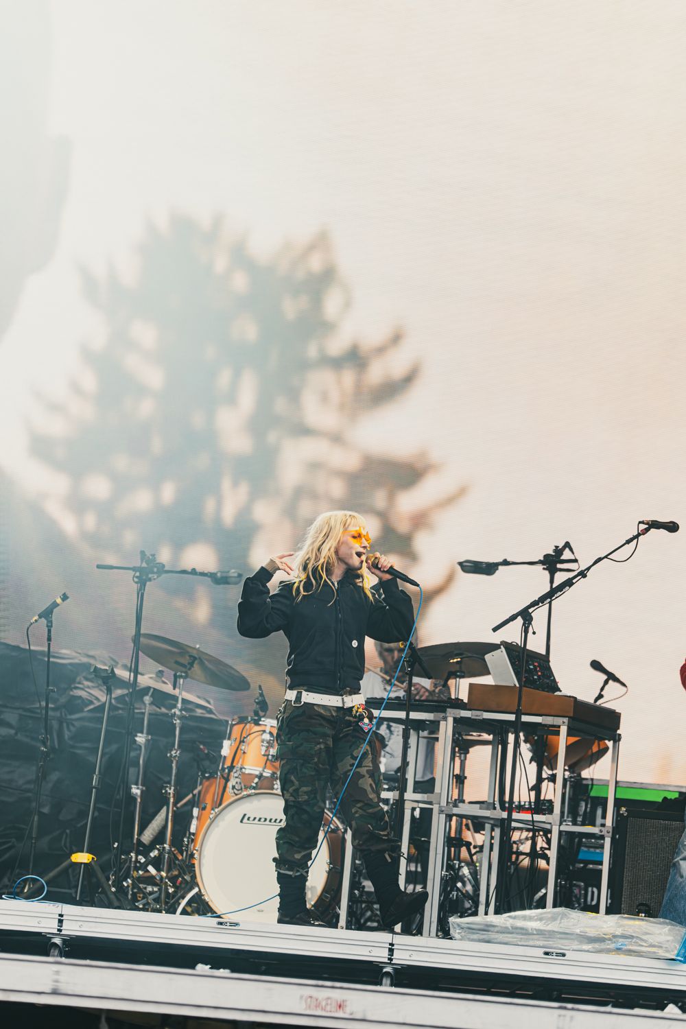 Blonde rock singer performs on outdoor stage with drum kit and misty atmosphere in background.