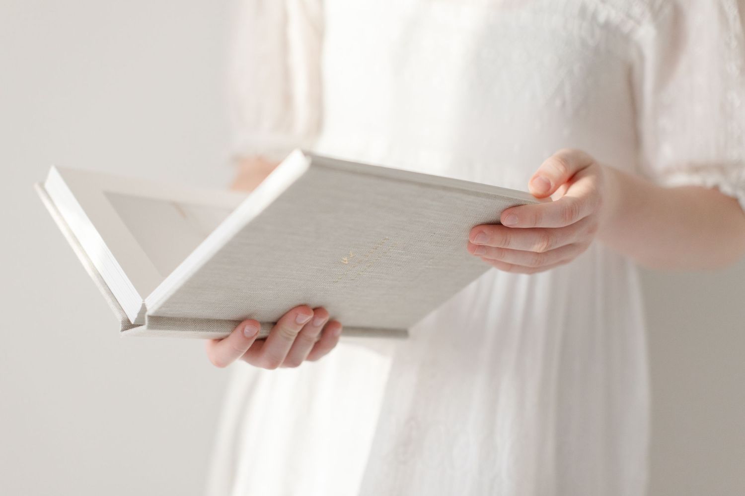 Woman in white dress holding an open white hardcover book with both hands against a light background.