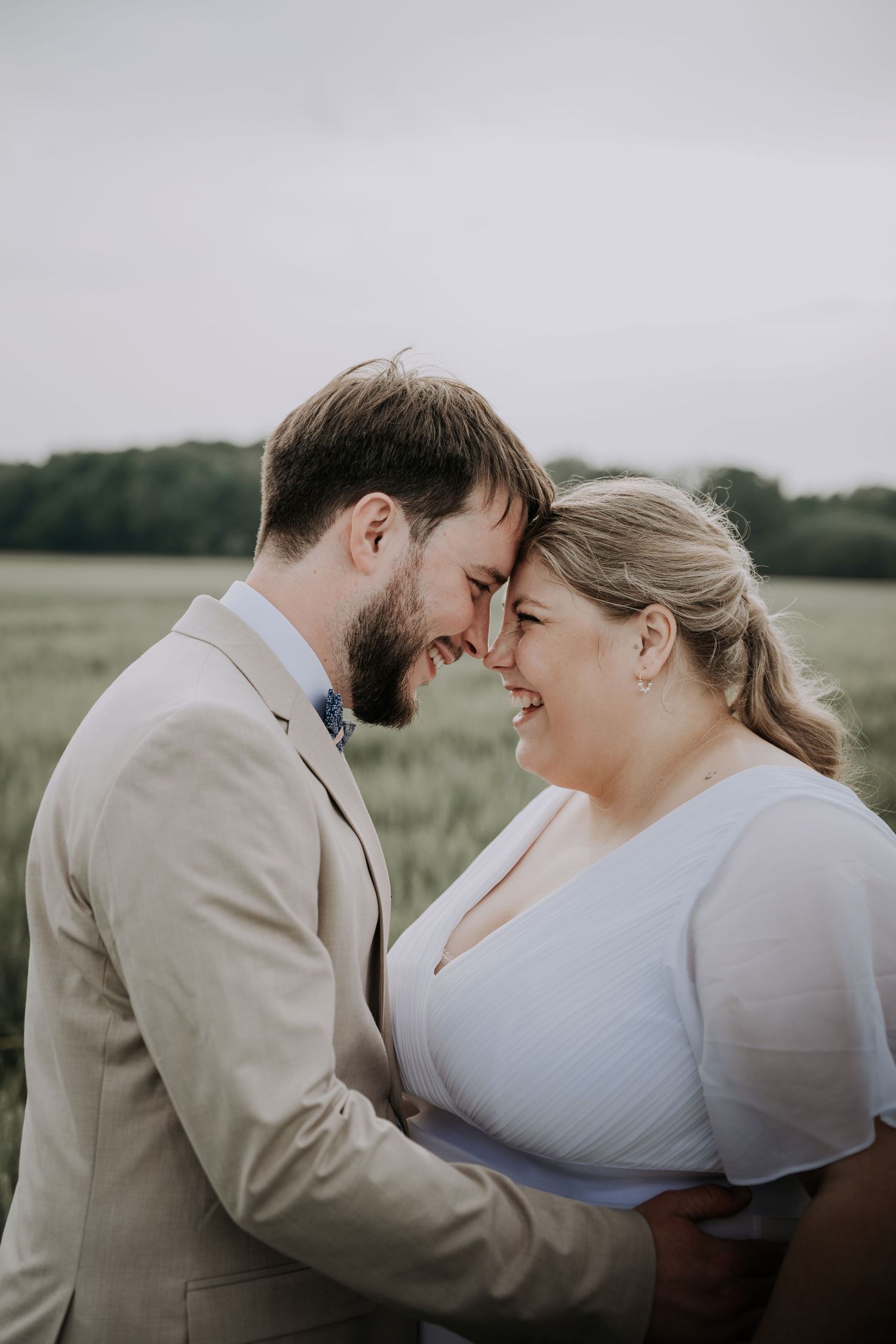 A couple in wedding attire share a romantic forehead touch in a green field, smiling joyfully on their wedding day.