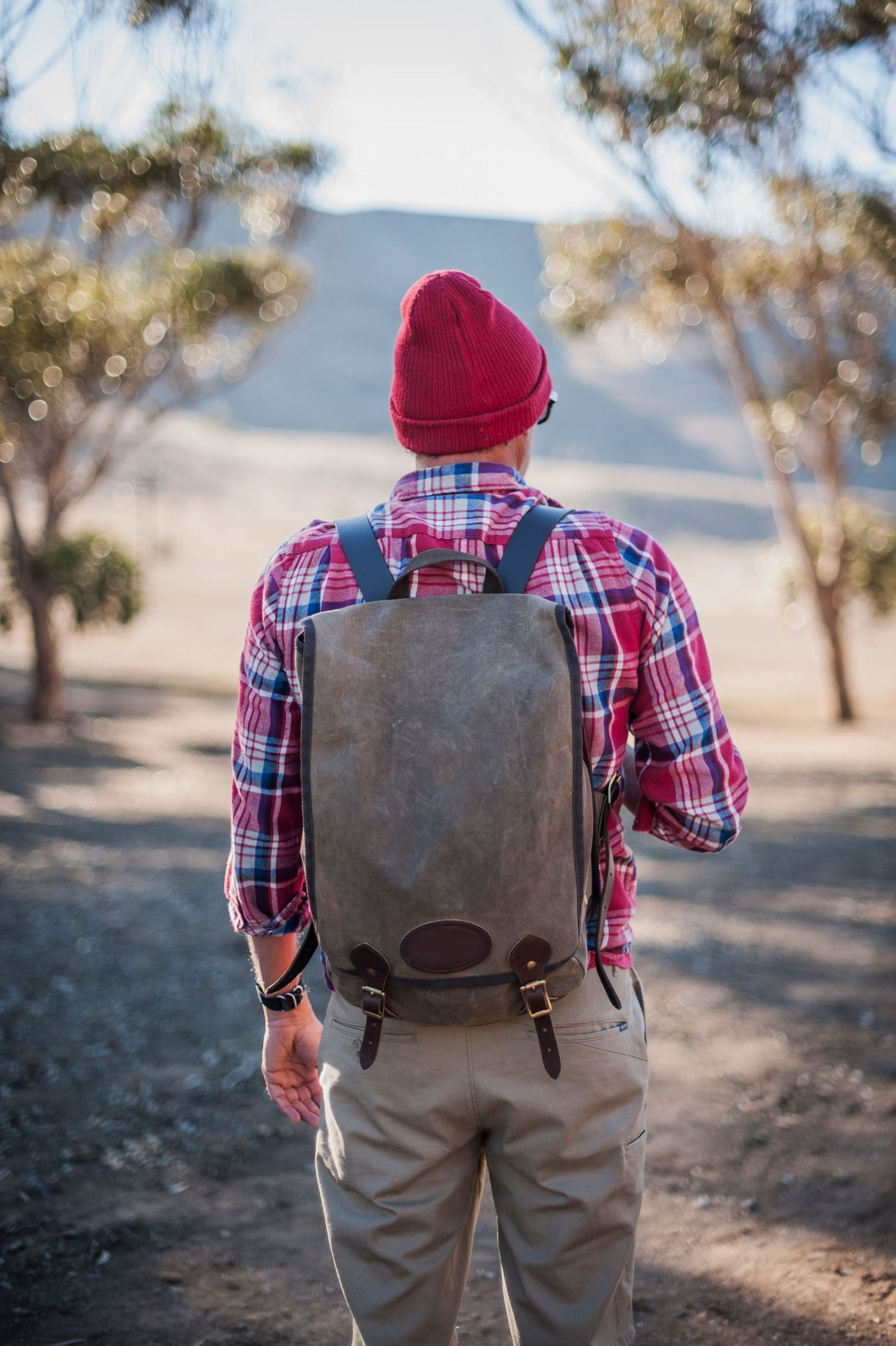 Hiker wearing red plaid shirt, beanie, and canvas backpack with leather straps, standing on a dirt trail surrounded by trees.