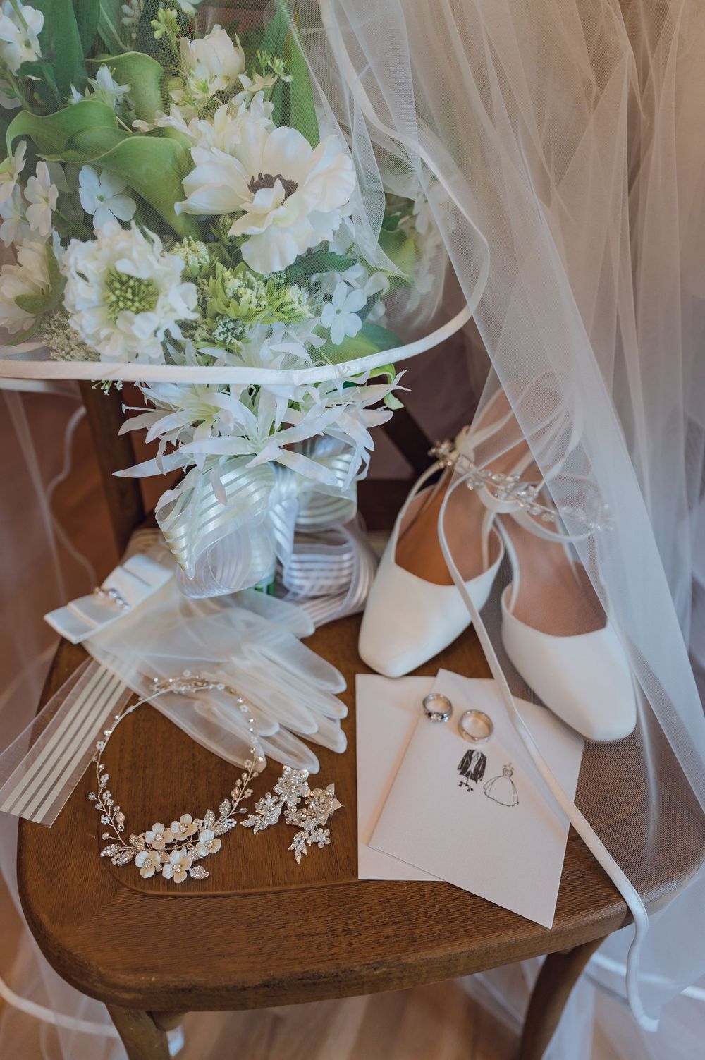 Bridal accessories flatlay featuring white heels, floral crown, gloves, earrings, and a white flower bouquet on a wooden table.