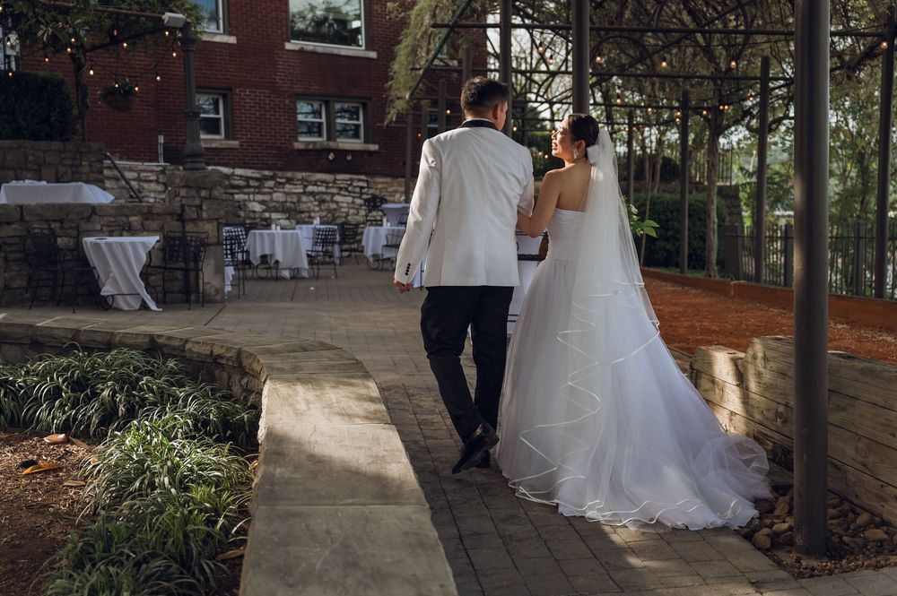 A bride in a white gown and groom in a white jacket walk hand in hand along a garden path at an outdoor wedding venue.