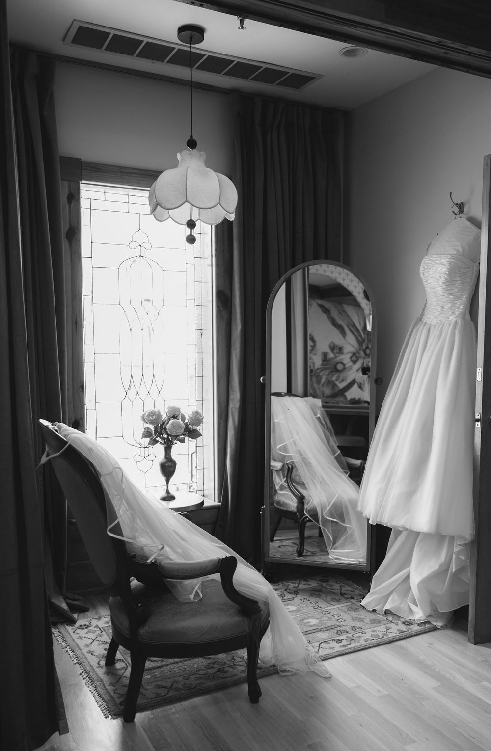 Black and white photo of a wedding dress hanging near an arched mirror, with a veil draped over a vintage chair by a stained glass window.