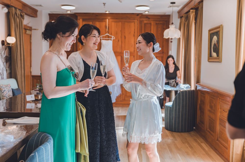 Three women in elegant dresses laugh and chat indoors, with a wedding gown hanging in the background.