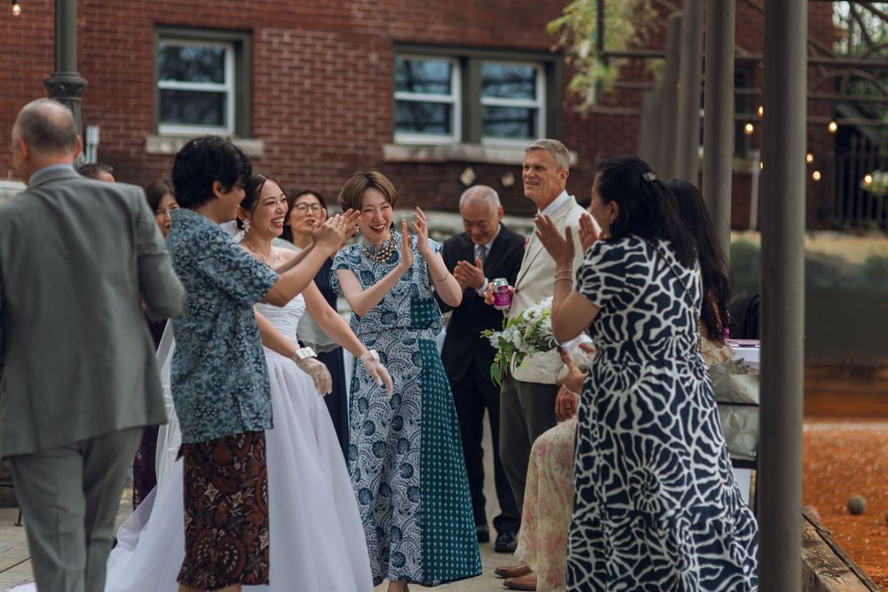 Wedding guests celebrate and applaud the newlyweds outdoors near a brick building with string lights.