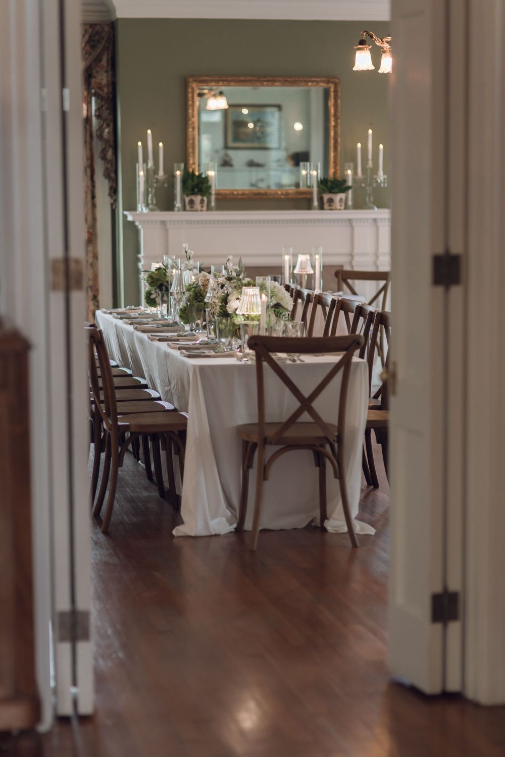 Elegant wedding reception dining room with long white tablecloth, wooden cross-back chairs, candles, and floral centerpieces.