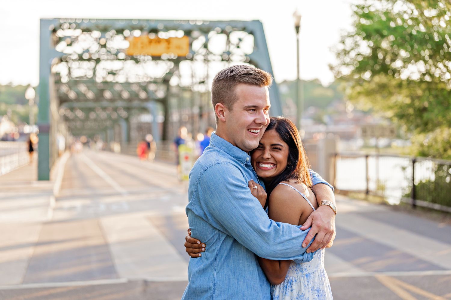 Happy newly engaged couple embracing and smiling on a historic bridge at golden hour.