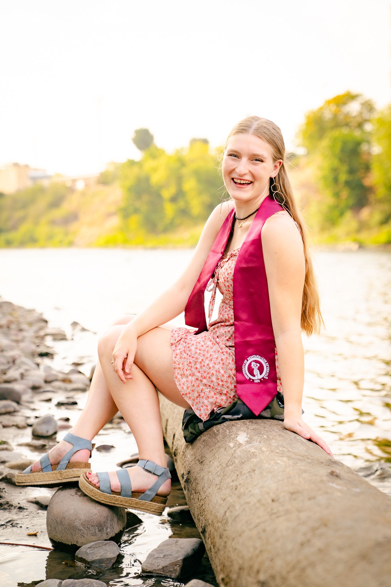 Smiling young woman in floral dress and maroon graduation stole poses outdoors in golden sunset light.