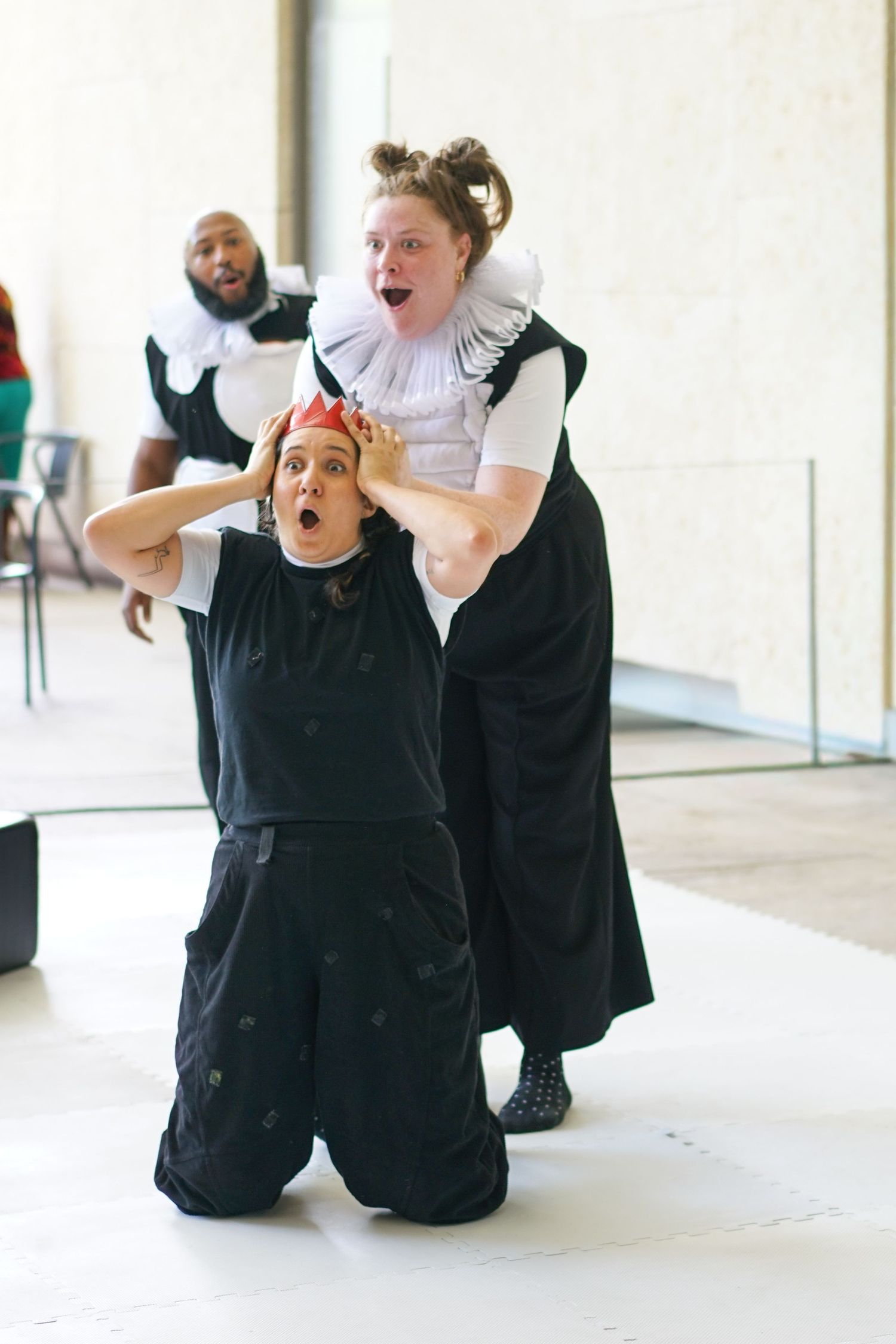 Two performers in black and white costumes act dramatically on stage, one kneeling with hands on head wearing a crown.