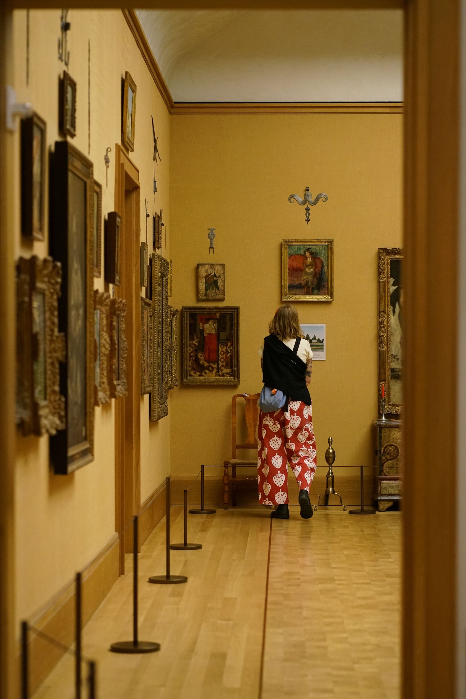 Woman in red floral skirt viewing paintings in a golden-walled art museum gallery with wooden floors.
