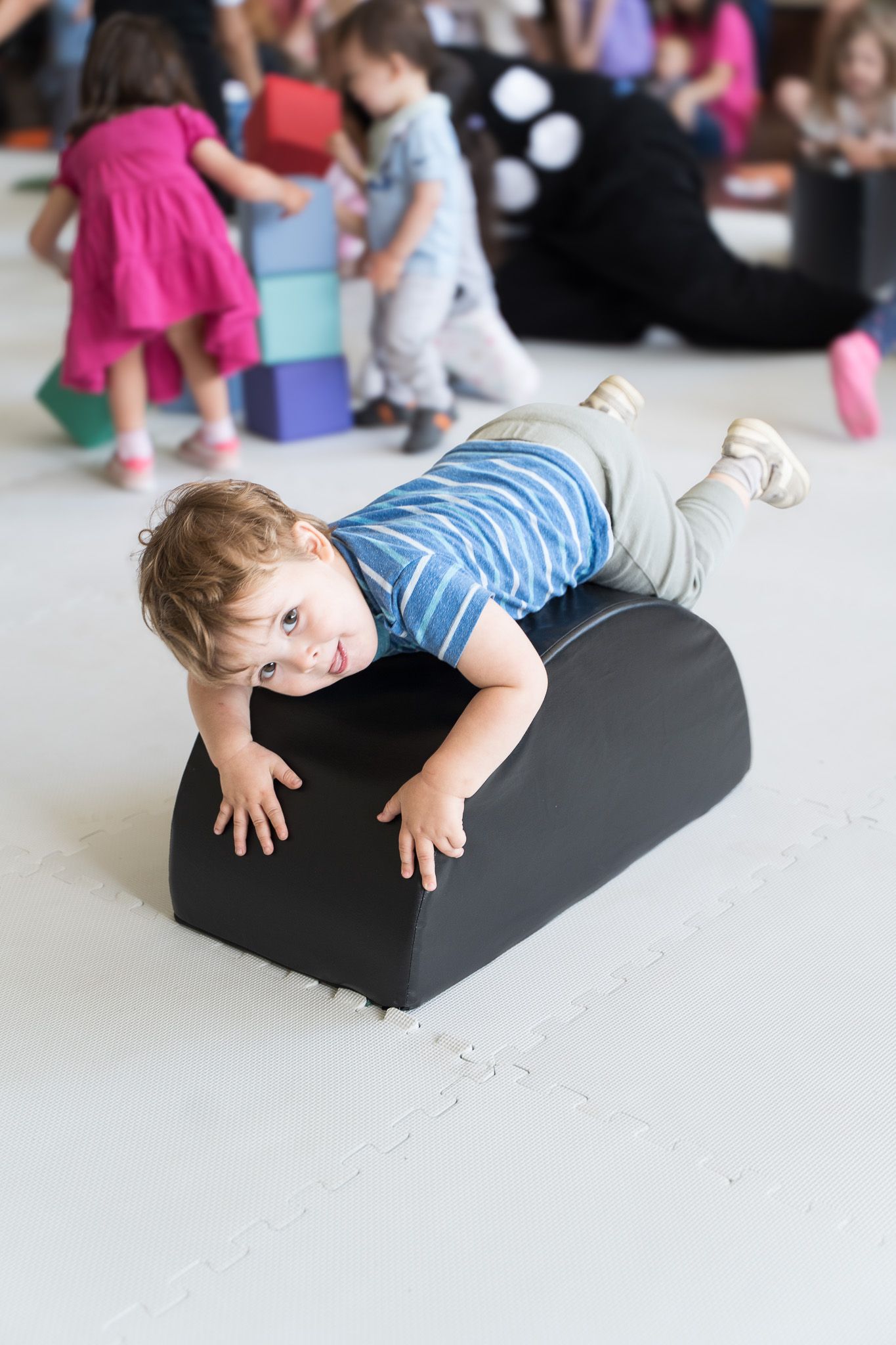 Toddler in striped shirt lying on black foam bolster during children's indoor play session.