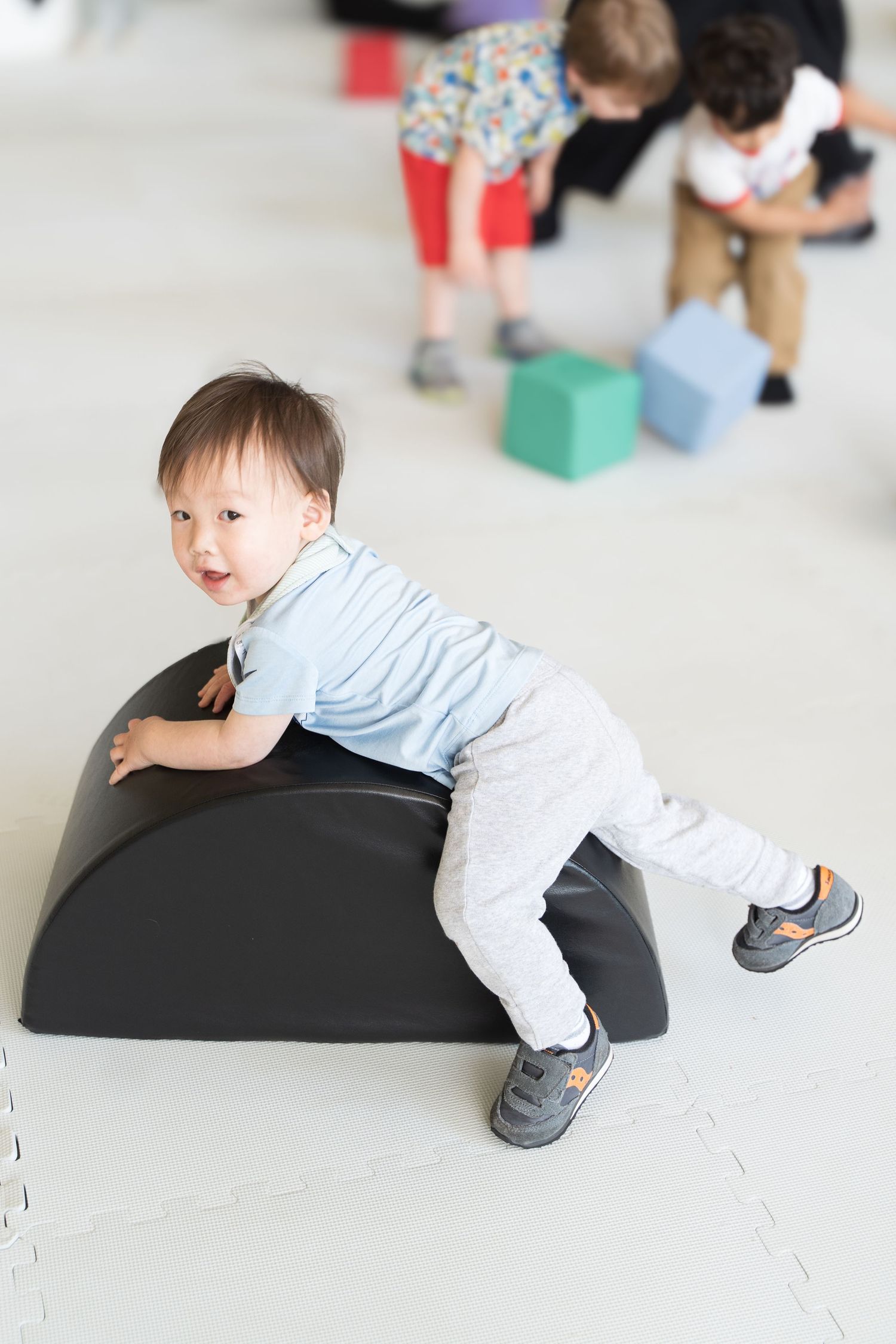Toddler boy climbing on a black foam balance rocker in a bright playroom with other children playing in the background.