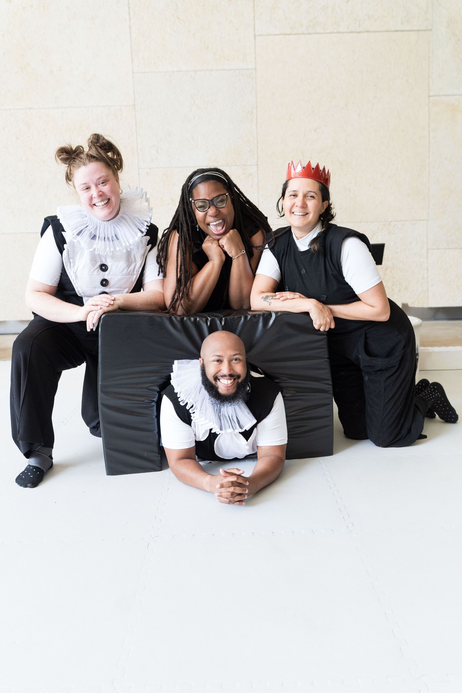 Four performers in black and white costumes pose together smiling, one wearing a paper crown, in a bright indoor space.