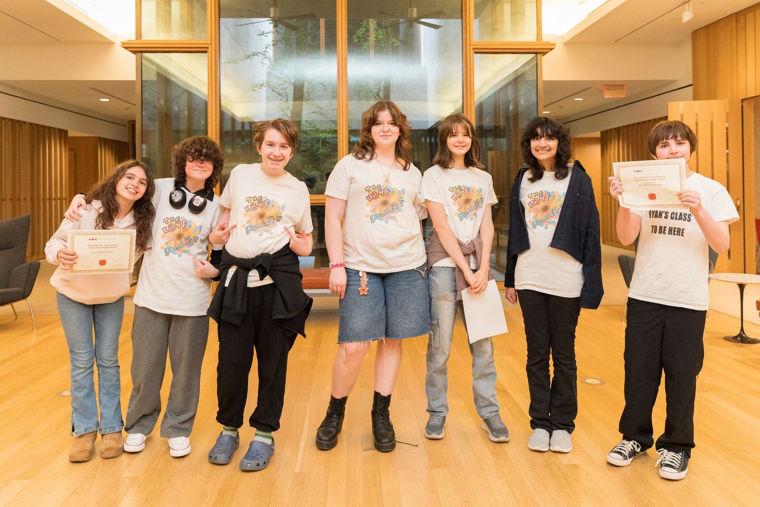 Seven students in matching white t-shirts pose together smiling in a modern building lobby.