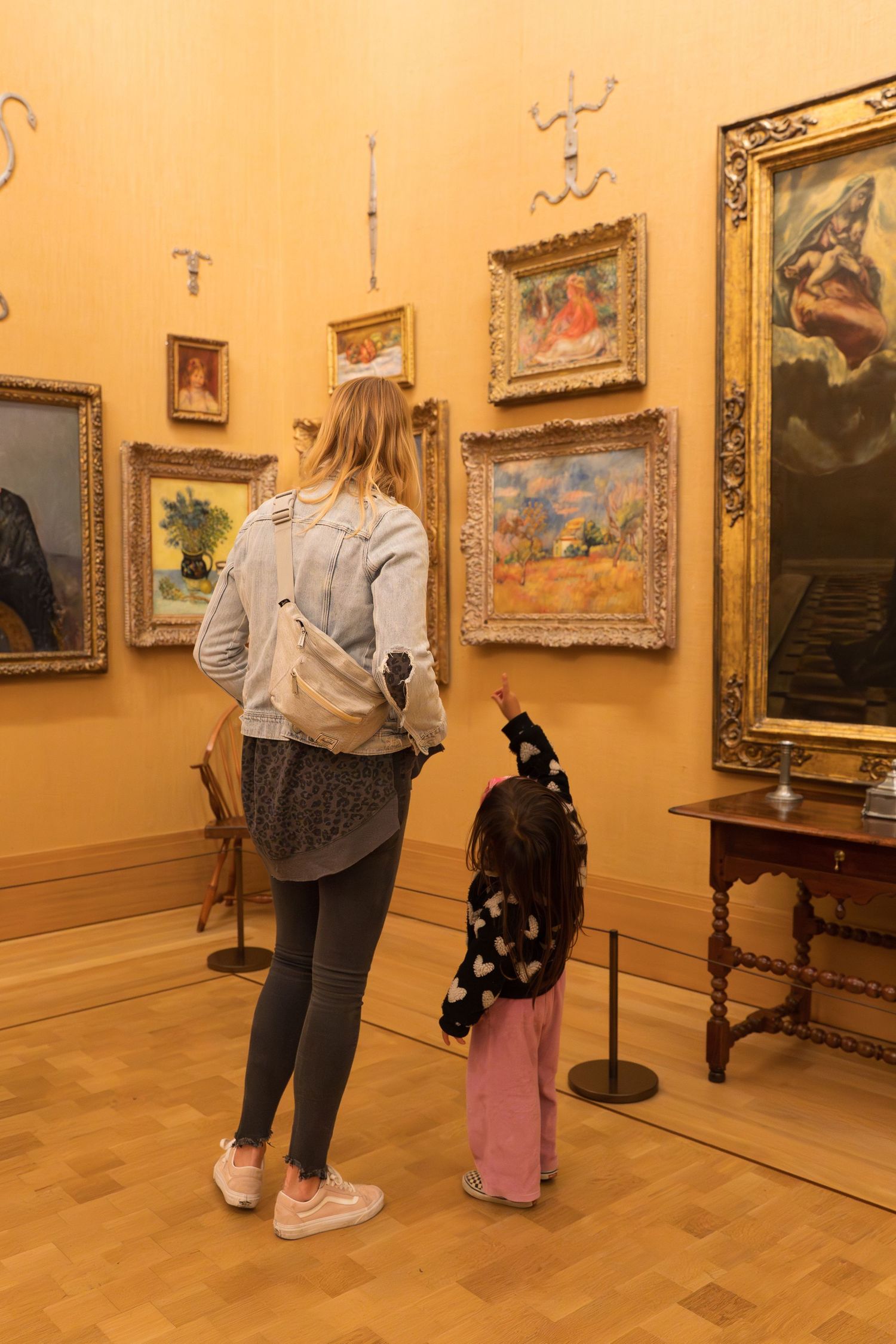 Mother and young child viewing classic paintings on yellow walls in an art museum gallery.