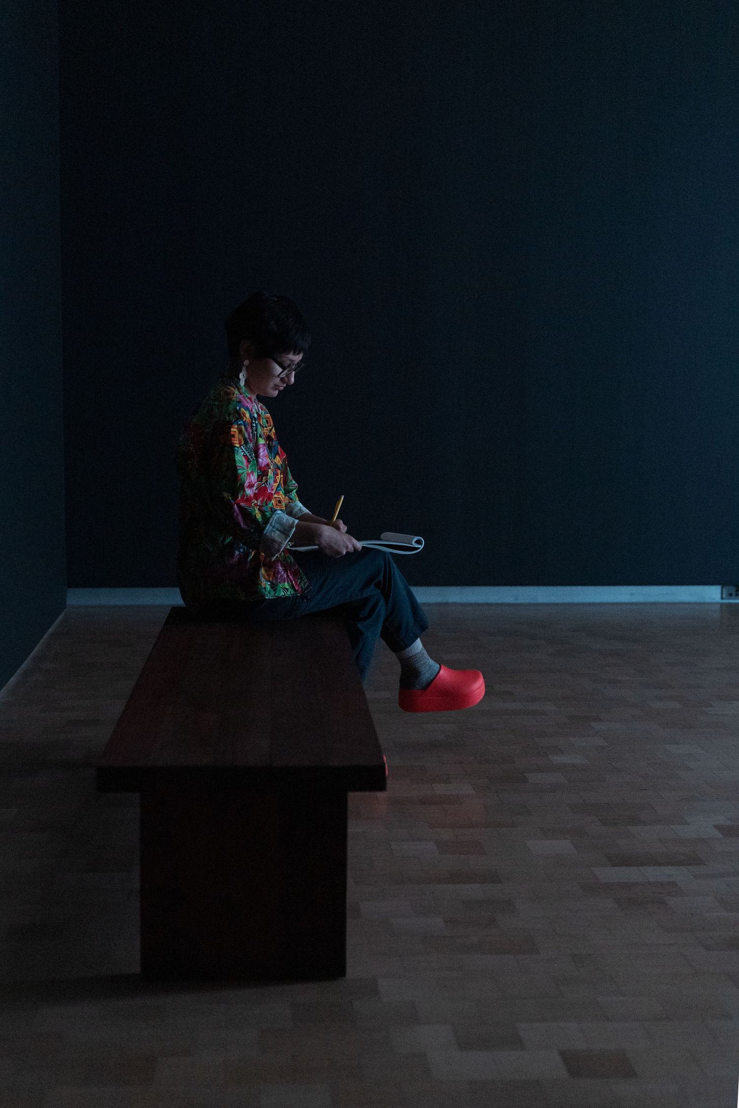 Person sitting on a dark wooden bench writing in a notebook, wearing a colorful shirt and red shoes in a dimly lit room.