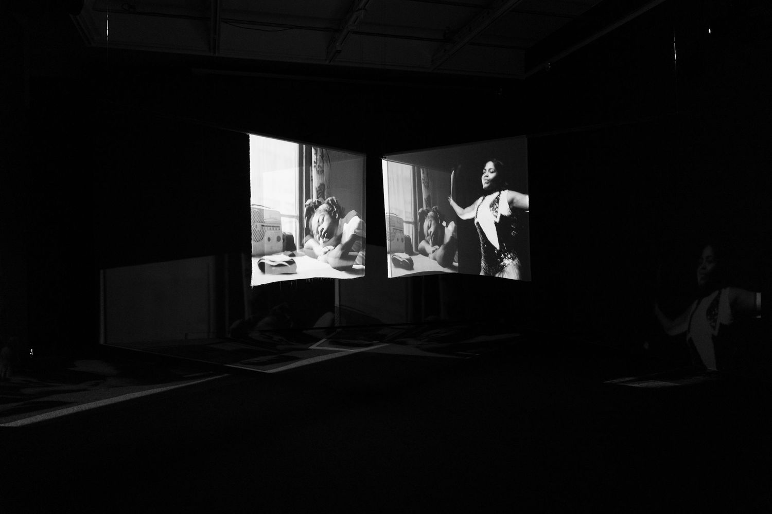 Dark room with two projected black-and-white film screens showing vintage scenes with people, in an art gallery setting.