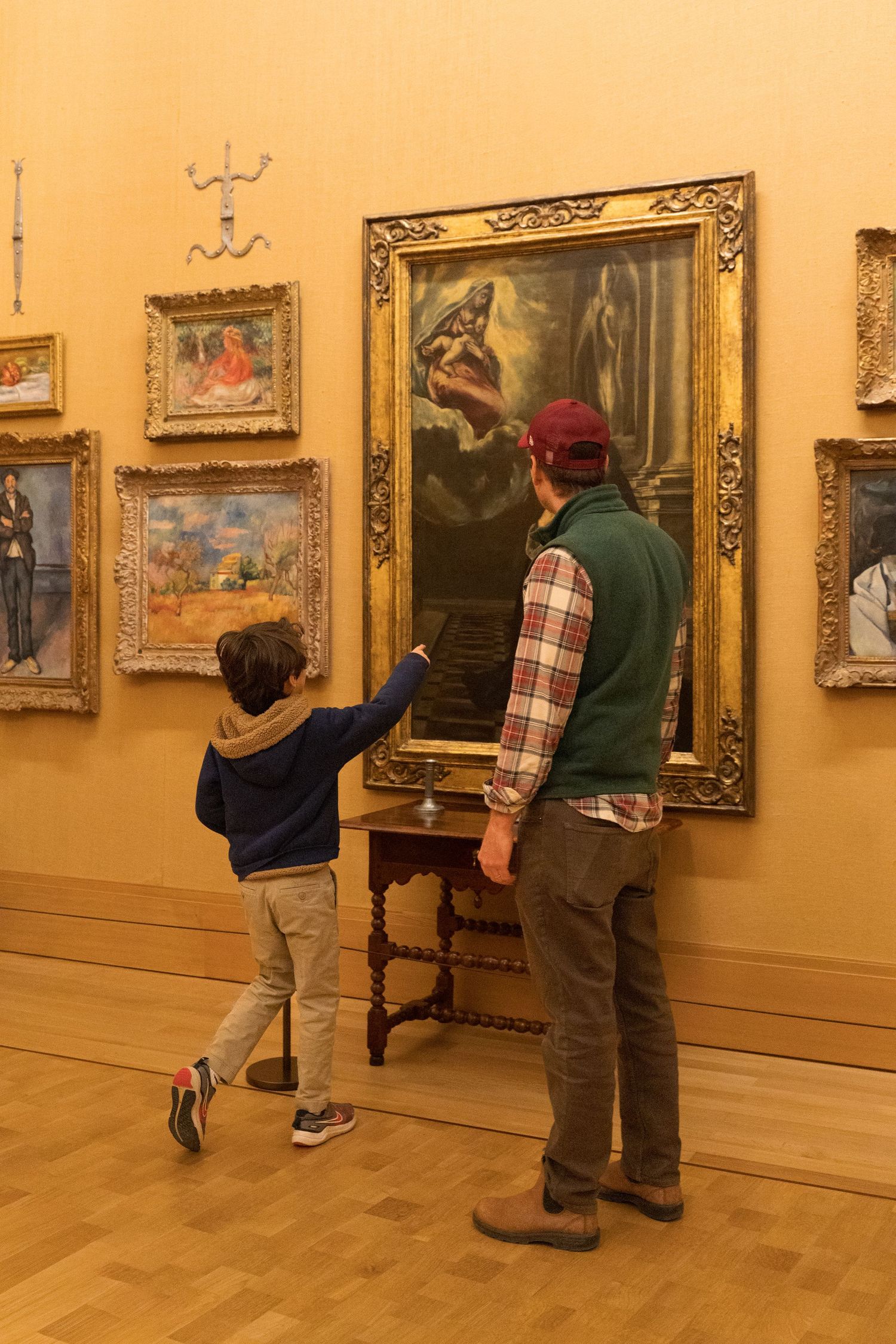 Adult and child viewing classical paintings in a museum gallery with ornate gold frames on warm yellow walls.
