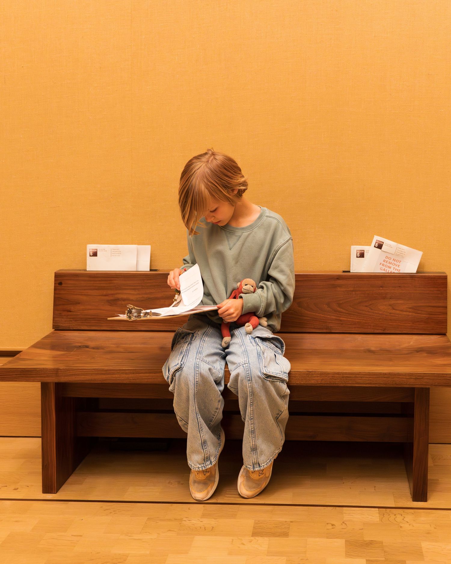 Young child sitting alone on wooden bench reading a booklet inside a museum gallery.